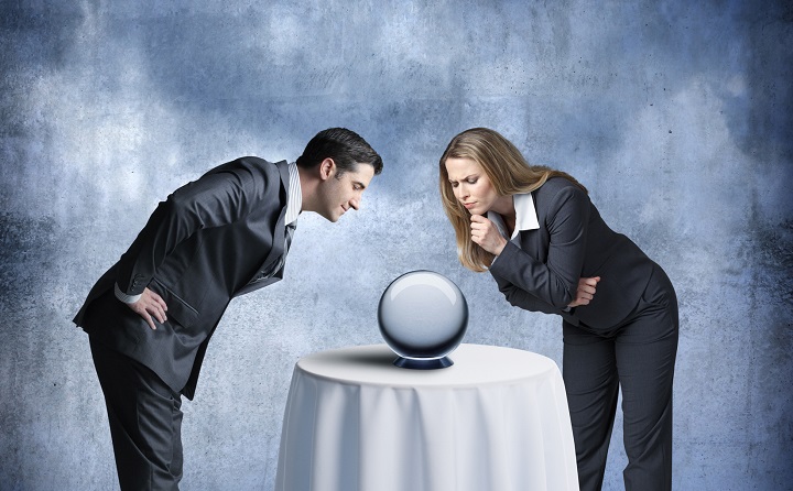 A businessman and a businesswoman look down at a crystal ball that sits on a tablecloth draped table in front of them.